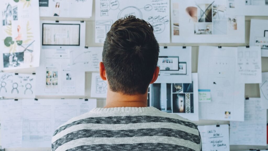 A person with short hair, viewed from behind, looks at a wall covered with various papers, diagrams, and photos deliberating using ai tools for research